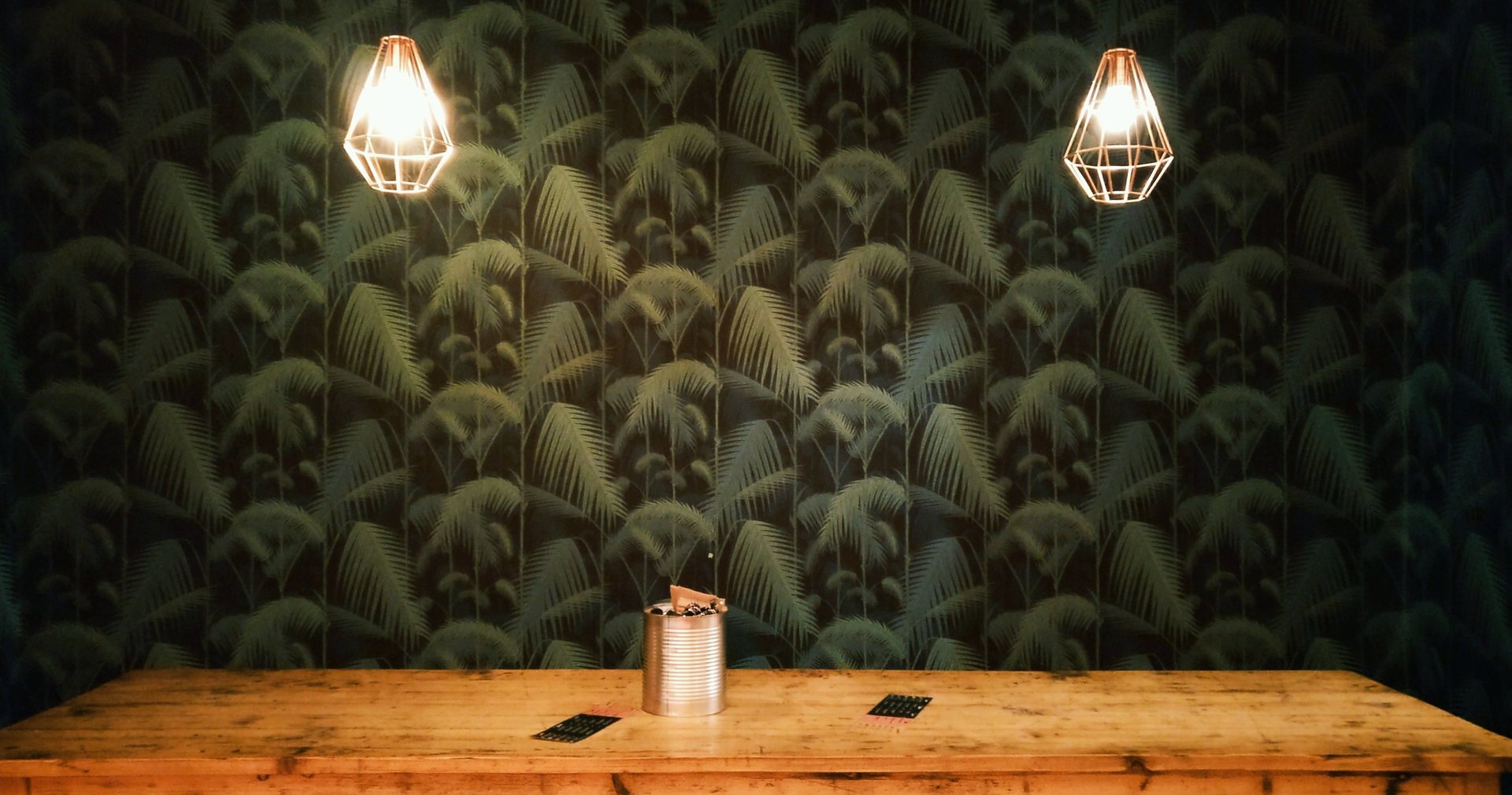 Dining space set against dark palm-frond wallpaper lit by pendant lamps